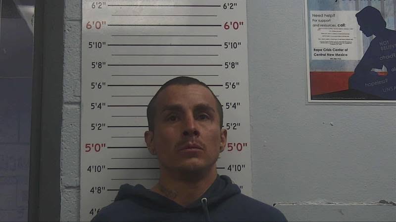 Police booking photo of 5'7" person facing forward with dark eyes, dark close cut hair and mustache, wearing dark blue hooded sweatshirt, showing neck tattoo.