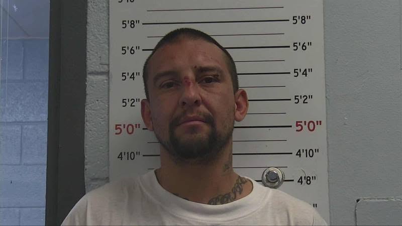 Police booking photo of 5'7" person facing forward with dark eyes, close cut dark hair and facial hair, abrasions on bridge of nose and above left eye, wearing white t-shirt and showing visible neck tattoos.
