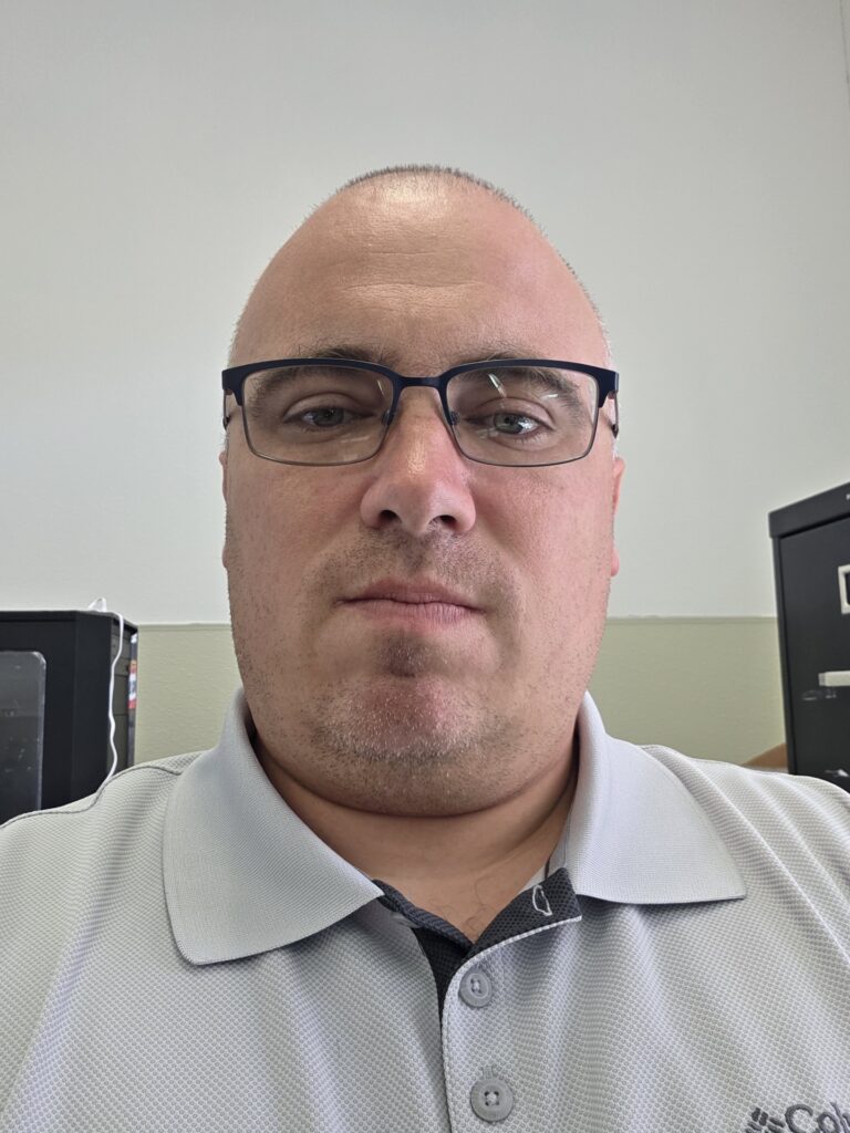 Person facing forward with blue eyes and shaved head, wearing black wireframe glasses and grey golf shirt.