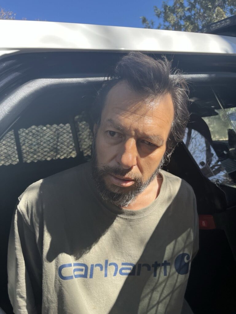 Person with dark eyes one slightly drooping, short dark hair, and greying facial hair facing forward with hands behind back at open door of police patrol unit wearing grey t-shirt showing navy blue brand logo.