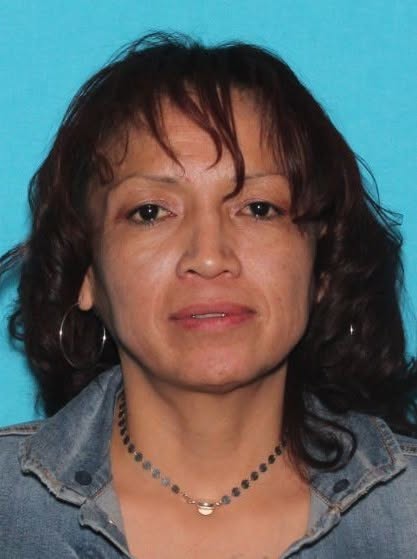 Person facing front with dark eyes and long hair with bangs, wearing silver hoop earring and silver chain necklace with light blue denim jacket.