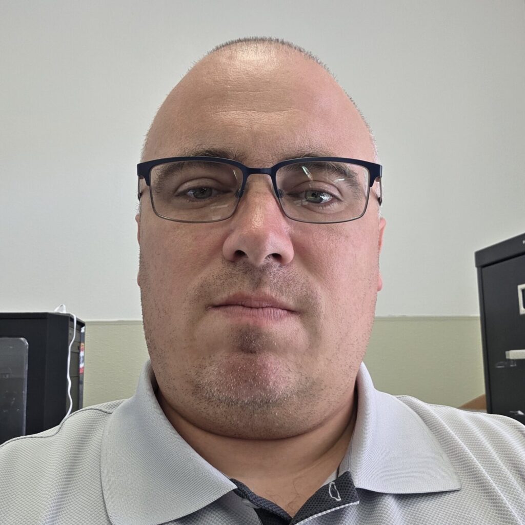 Person facing forward with blue eyes and shaved head, wearing black wireframe glasses and grey golf shirt.